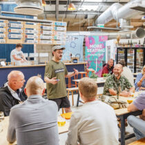 Guide in a Cloudwater t-shirt speaks to a group seated at tables inside the brewery taproom, with drinks, laughter, and a bar menu in the background.