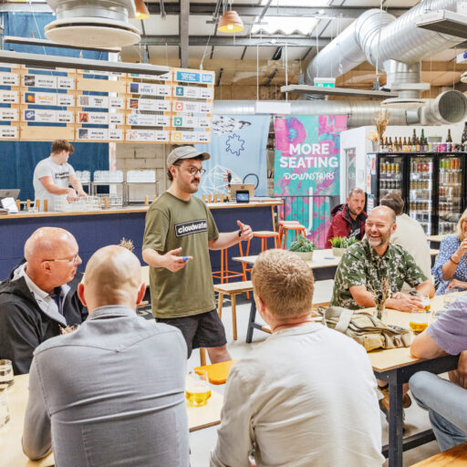 Guide in a Cloudwater t-shirt speaks to a group seated at tables inside the brewery taproom, with drinks, laughter, and a bar menu in the background.