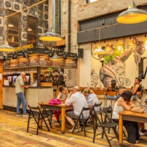 People enjoy drinks and conversation inside a lively bar with industrial décor, a central wine counter, and a large mural featuring a classical figure holding a head.