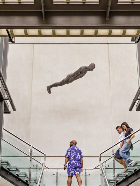 Man in a patterned purple outfit stands on a staircase looking up at a suspended human figure sculpture, while a couple descends nearby stairs inside a modern building.