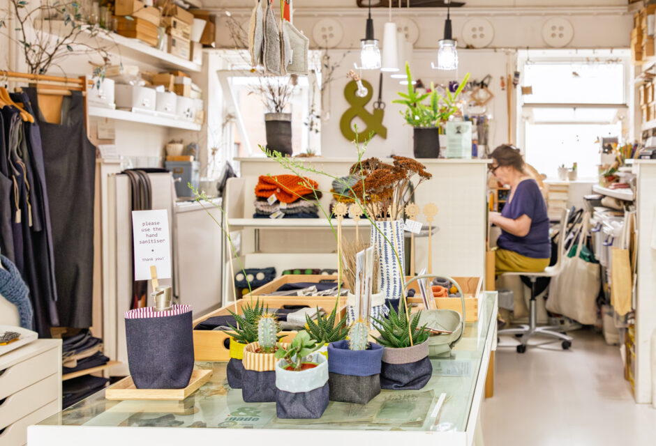 Creative studio filled with handmade crafts and plants, with a woman working at a desk in the background surrounded by materials.