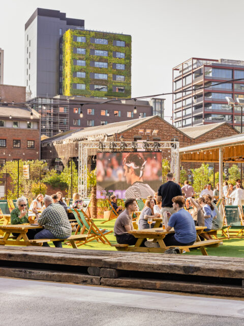 Outdoor social space with people at picnic tables, drinks in hand, and a large screen in the background showing a broadcast.