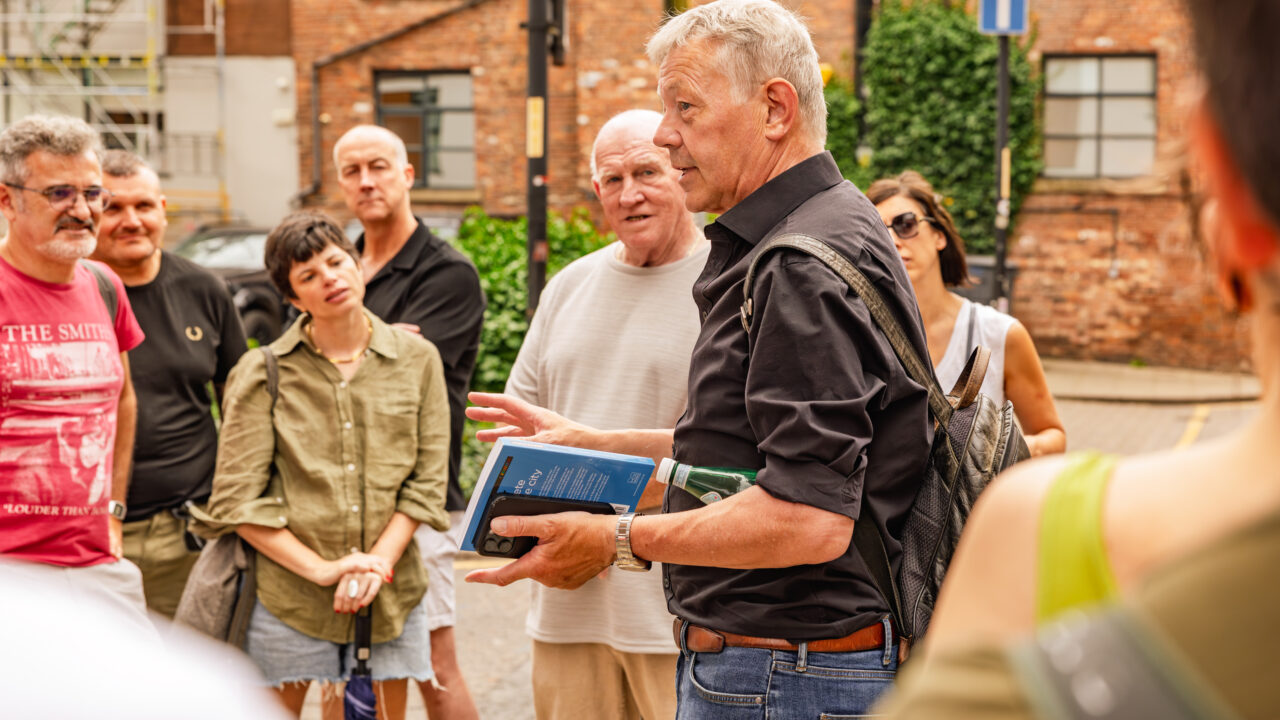 A male tour guide speaking to a group on a city street, holding a book while participants listen attentively.