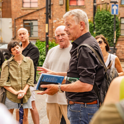A male tour guide speaking to a group on a city street, holding a book while participants listen attentively.