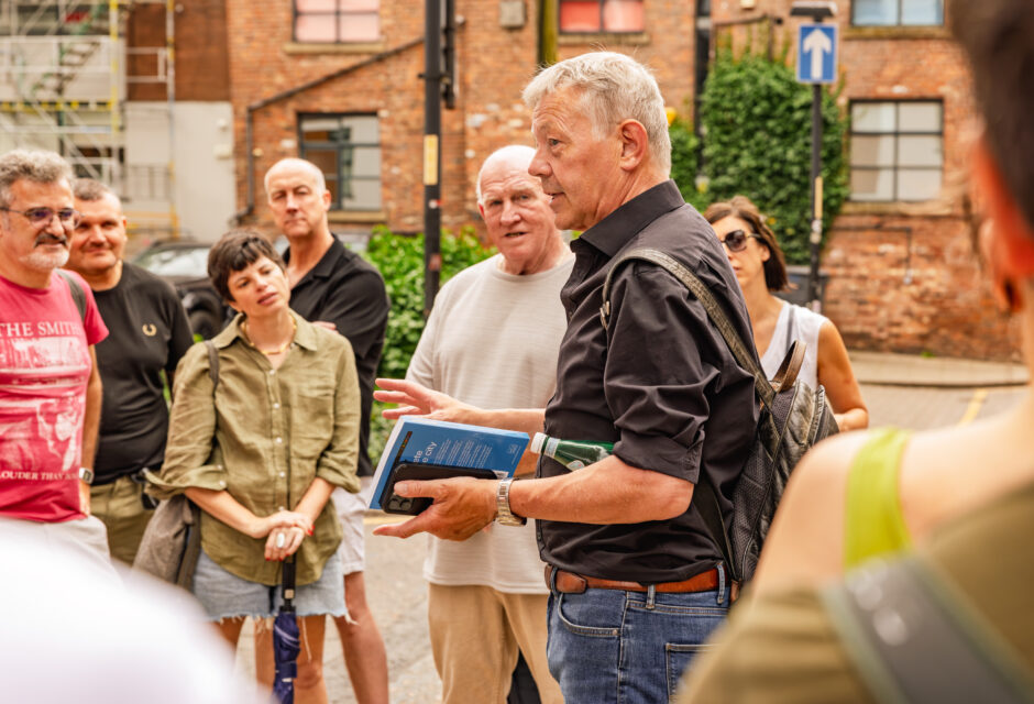A male tour guide speaking to a group on a city street, holding a book while participants listen attentively.