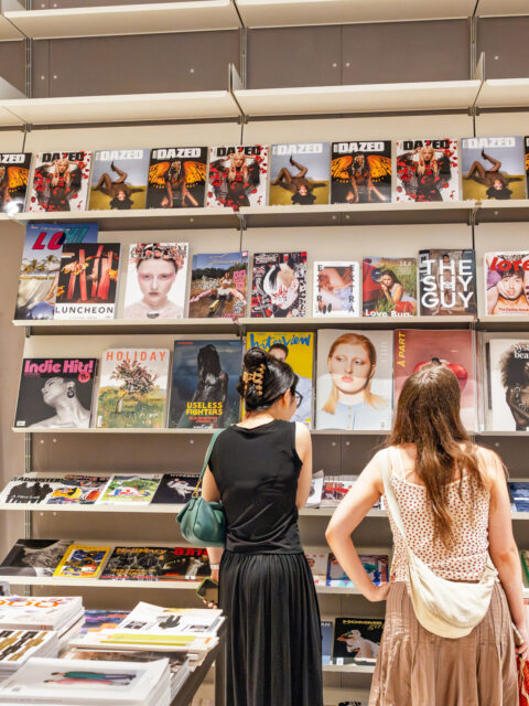 Two women browse a wall of art and fashion magazines in a modern shop, with colourful covers including Dazed, Love, and Interview.