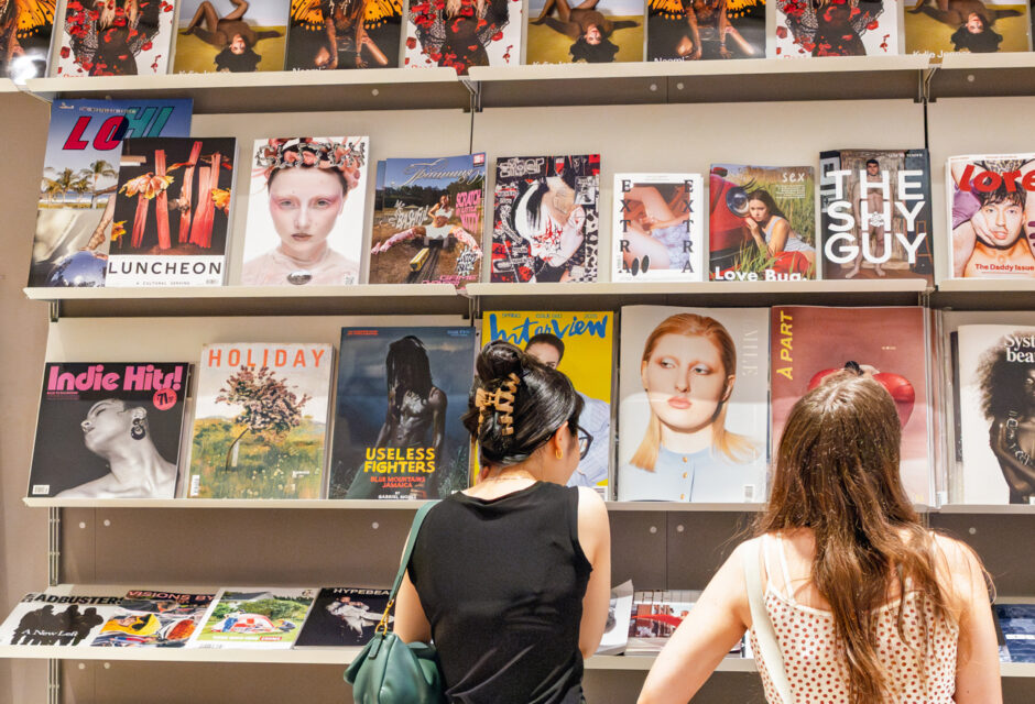 Two women browse a wall of art and fashion magazines in a modern shop, with colourful covers including Dazed, Love, and Interview.