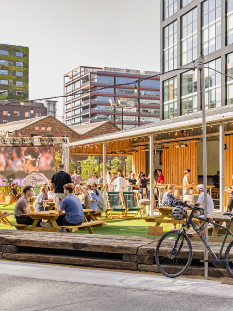 Outdoor social space with people at picnic tables, drinks in hand, and a large screen in the background showing a broadcast.