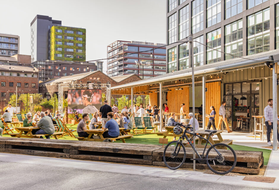 Outdoor social space with people at picnic tables, drinks in hand, and a large screen in the background showing a broadcast.