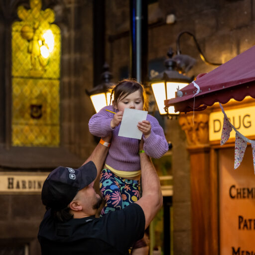 Child holding paper and pencil being held up by an adult in the Victorian Street at Abbey House Museum