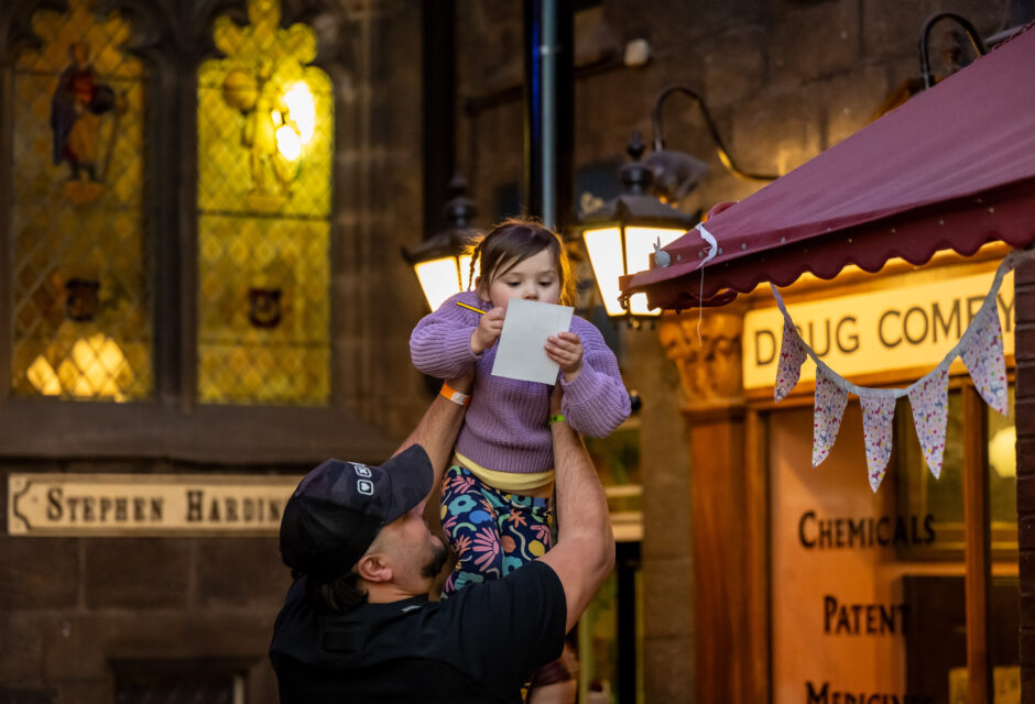 Child holding paper and pencil being held up by an adult in the Victorian Street at Abbey House Museum